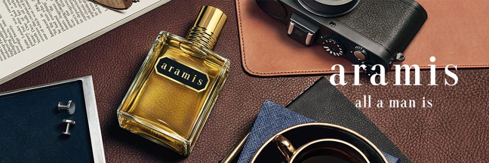 Rappresentazione piatta di un'elegante composizione con un flacone di profumo Aramis, una macchina fotografica, gemelli, un libro aperto, un taccuino e una tazza di caffè su una superficie marrone strutturata. Il testo recita: "Aramis è tutto ciò che un uomo è".