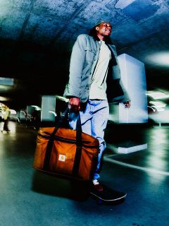 Uomo che va in skateboard in un parcheggio, con una grande borsa Carhartt marrone, una giacca verde e jeans sotto una luce artificiale.