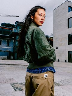 Una donna con lunghi capelli scuri, giacca a vento verde e pantaloni marrone chiaro si trova in un ambiente urbano all'aperto e guarda oltre la sua spalla.