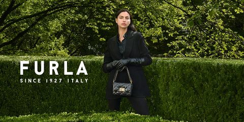 Una donna è in piedi davanti a una vegetazione lussureggiante. Indossa un abito nero e guanti e tiene in mano una borsa a mano nera trapuntata. A sinistra si legge il testo "Furla Since 1927 Italy".