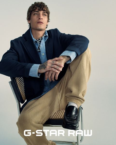 Una persona è seduta su una sedia e indossa un blazer scuro, una camicia di jeans e pantaloni beige. Ha dei tatuaggi sulla mano e guarda in alto. La persona indossa anche delle scarpe basse e in basso nell'immagine c'è il testo "G-STAR RAW".