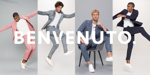 Quattro uomini in eleganti completi e scarpe da ginnastica bianche sono visibili su uno sfondo luminoso. Due saltano energicamente, uno è sdraiato su una sedia e uno posa. La parola "BENVENUTO" è sovrapposta all'immagine.