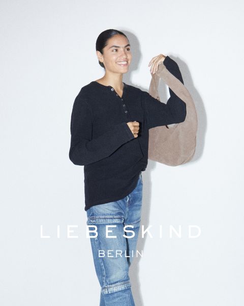 Una donna sorridente con una camicia nera a maniche lunghe e jeans indossa una borsa marrone chiaro a tracolla. Il testo "Liebeskind Berlin" appare nella parte inferiore dell'immagine.