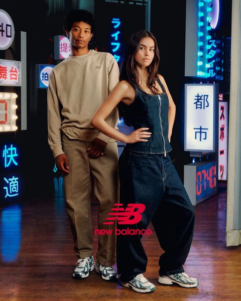 Un uomo e una donna posano davanti a insegne al neon. L'uomo indossa una tuta beige, la donna una salopette di jeans. Entrambi indossano scarpe da ginnastica New Balance. La scena è illuminata da diverse luci al neon blu e rosse.