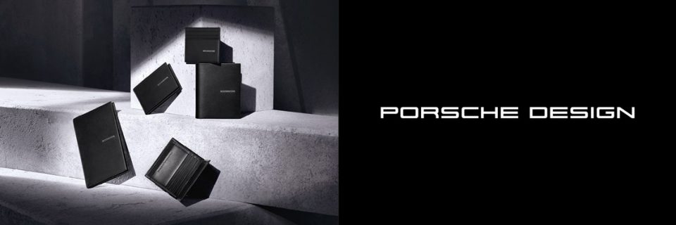 Una collezione di accessori in pelle nera di Porsche Design, tra cui portafogli e porta carte, è presentata elegantemente su superfici in cemento, proiettando ombre delicate. Il logo Porsche Design è ben visibile sulla destra.