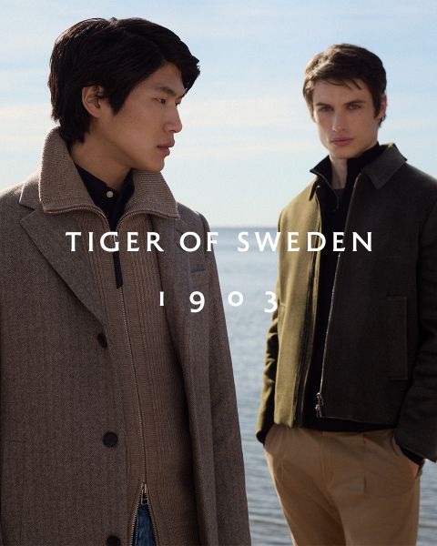 Due uomini in cappotti eleganti sono in piedi all'aperto vicino all'acqua sotto un cielo limpido. Il testo "TIGER OF SWEDEN 1903" è centrato nell'immagine.