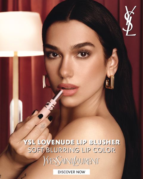 Una donna che si applica un rossetto rosa e indossa orecchini a cerchio dorati, davanti a tende rosse. Il testo pubblicizza lo „YSL Lovenuede Lip Blusher“, un rossetto dalla sfumatura morbida.