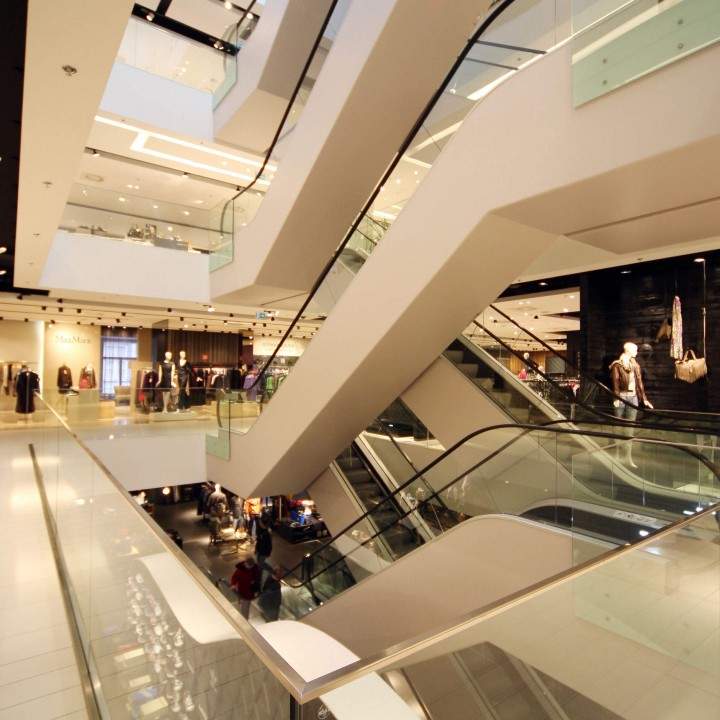 Un moderno centro commerciale con diversi piani e scale mobili. Lo spazio è ben illuminato e presenta negozi con vetrine su vari livelli. Ovunque si notano ringhiere in vetro ed eleganti elementi di design architettonico.