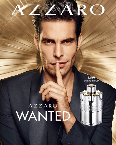 Un uomo in giacca e cravatta si porta un dito alle labbra in un gesto di pacificazione. Davanti a lui c'è una bottiglia di Azzaro Wanted Eau de Parfum. Lo sfondo mostra un design metallico circolare. Il testo recita "Azzaro" e "Wanted".