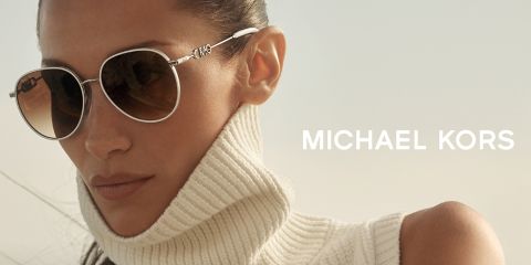 Una persona con occhiali da sole rotondi e un dolcevita bianco guarda di lato. Lo sfondo è leggermente sfocato e a destra si vede il nome "Michael Kors" in caratteri bianchi.