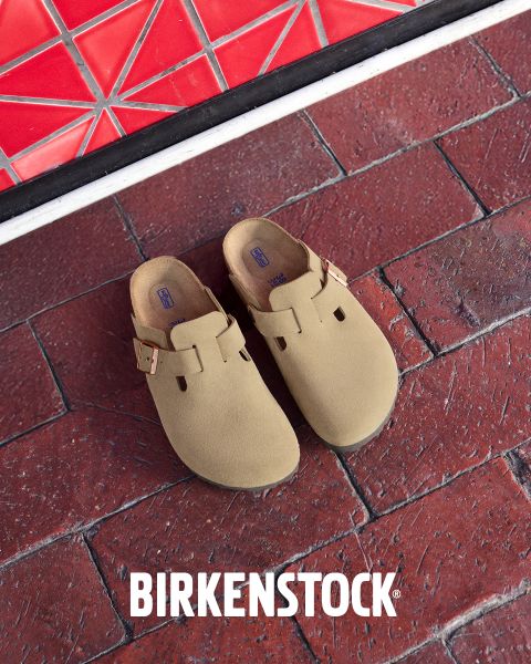 Un paio di zoccoli Birkenstock beige con cinturini con fibbia su un pavimento di mattoni rossi accanto a una parete piastrellata rossa geometrica. Il logo bianco Birkenstock si trova nella parte inferiore dell'immagine.