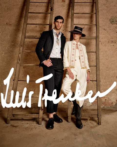 Un uomo e una donna in abiti eleganti di ispirazione vintage sono in piedi tra due scale di legno davanti a un muro rustico. L'uomo indossa un abito a righe scure, la donna un abito color crema con cappello e stivali. "Luis Trenker" è scritto in grassetto.