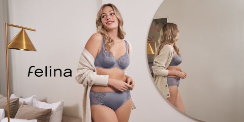 Una donna in biancheria intima grigia sorride in una stanza con uno specchio rotondo. Indossa un cardigan beige e guarda lontano dallo specchio. Accanto a lei c'è una moderna lampada da terra dorata. A sinistra si vede la parola "Felina".