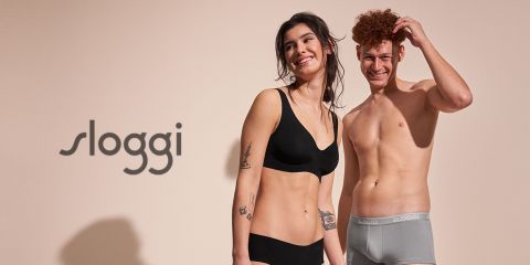 Una donna sorridente e un uomo in biancheria intima Sloggi sono in piedi davanti a uno sfondo neutro con il logo del marchio sul lato sinistro. La donna indossa un completo nero e l'uomo uno slip grigio.
