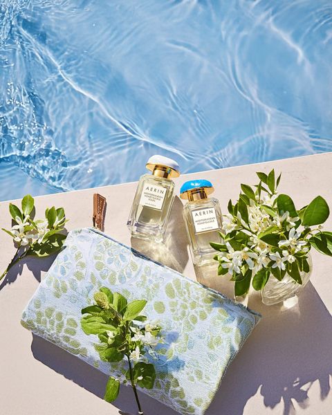 Due flaconi di profumo, una pochette decorata con fiori e piccoli vasi con fiori bianchi sono disposti su una mensola a bordo piscina, vicino all'acqua limpida e blu.