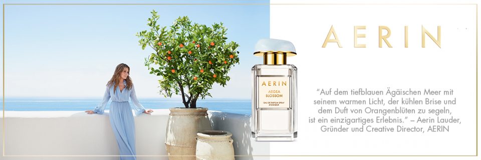 Una donna in un abito azzurro è in piedi su una terrazza con vista sul mare accanto a un albero di arance. In primo piano c'è una bottiglia di profumo Aerin, su cui il marchio è chiaramente visibile. Accanto alla bottiglia si vede un testo in tedesco.