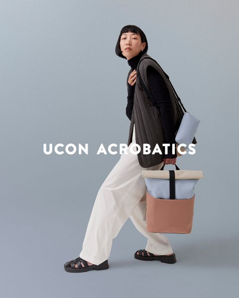 Una persona con un taglio di capelli a caschetto posa davanti a uno sfondo sobrio. Indossa un top nero, pantaloni bianchi e sandali e porta uno zaino alla moda. Sopra l'immagine c'è scritto "Ucon Acrobatics".