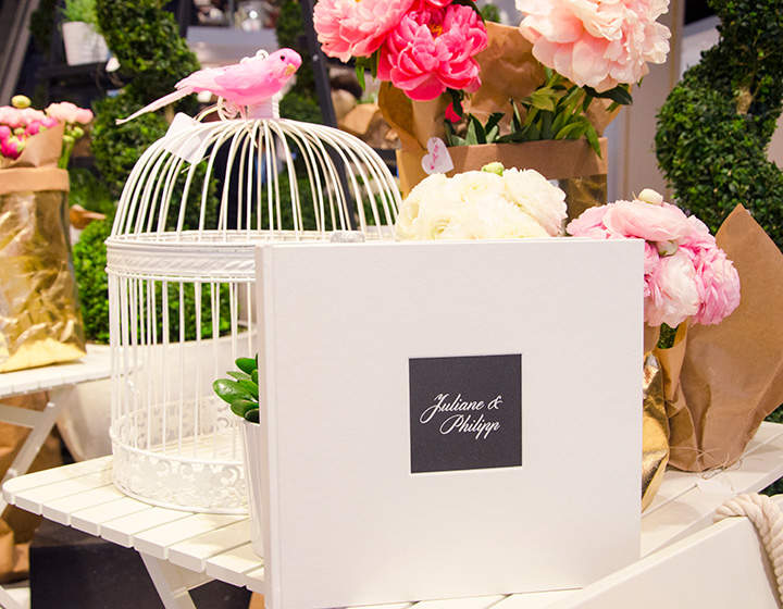 Un tavolo decorato con fiori rosa e bianchi, una gabbia per uccelli bianca decorata con un piccolo uccellino rosa e un libro bianco con un'etichetta nera con scritto "Juliane & Philipp".