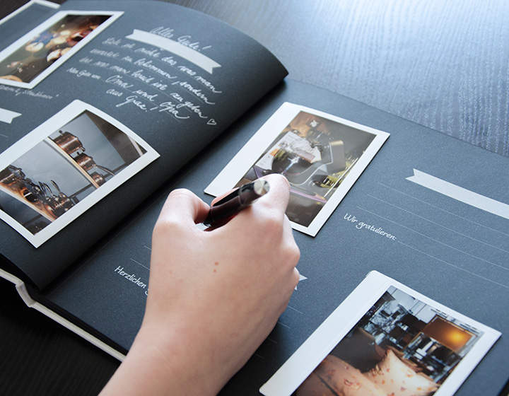 La mano di una persona tiene una penna e scrive in un album nero, sulle cui pagine sono attaccate delle foto. Il libro è aperto su un tavolo scuro. Le pagine hanno spazio per scrivere e diverse immagini con sfondo chiaro.
