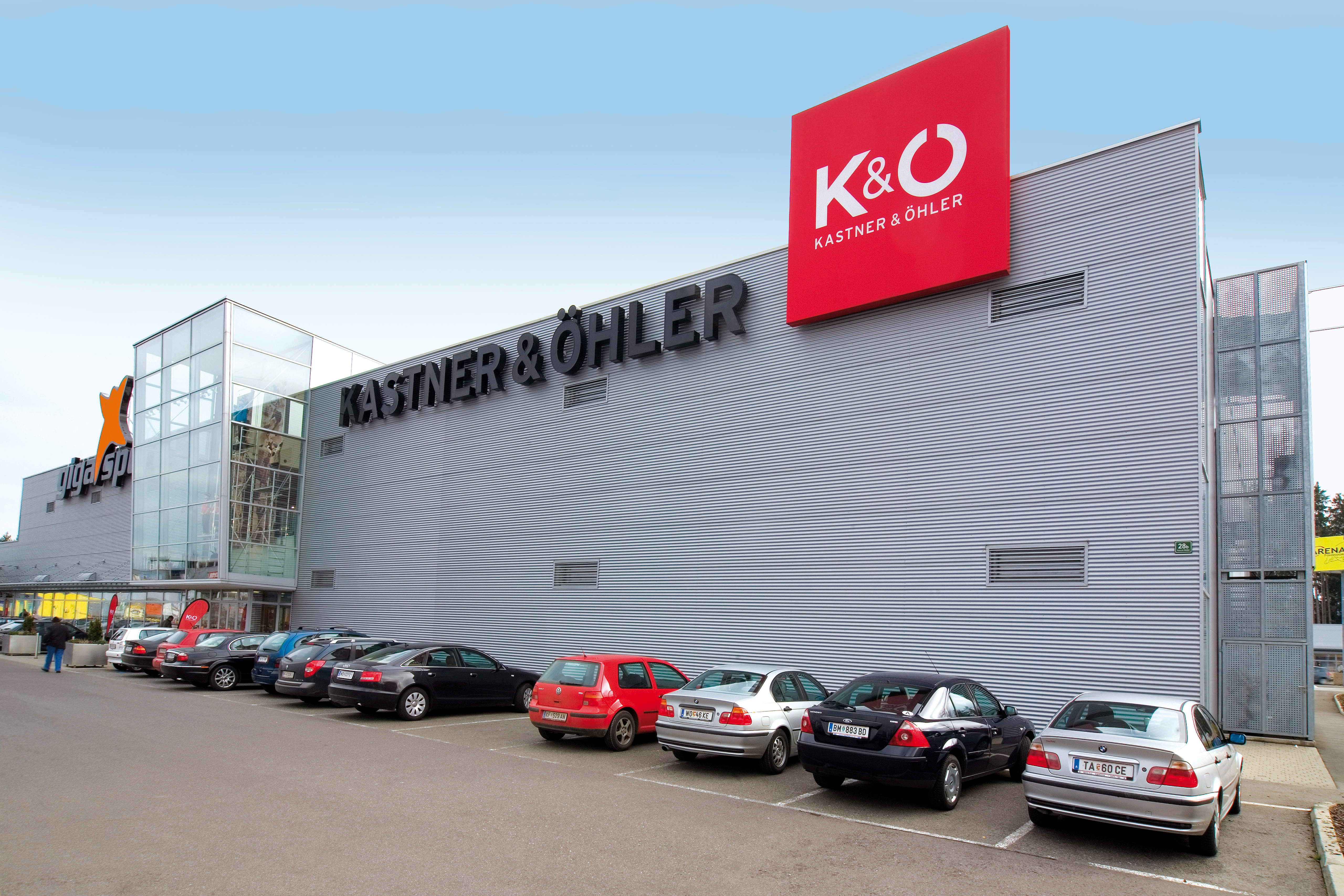 Un moderno edificio commerciale con la scritta e il logo "Kastner & Öhler" sulla facciata. Davanti all'edificio, che ha una grande superficie vetrata sul lato sinistro, diverse auto sono parcheggiate nel parcheggio. Il cielo è sereno.