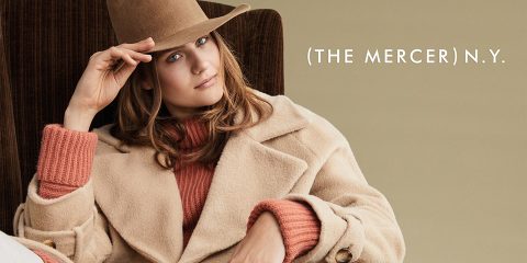 Una persona con un cappotto beige e un cappello è seduta su una poltrona marrone. Accanto a lei, su uno sfondo beige chiaro, si legge il testo "The Mercer N.Y.".