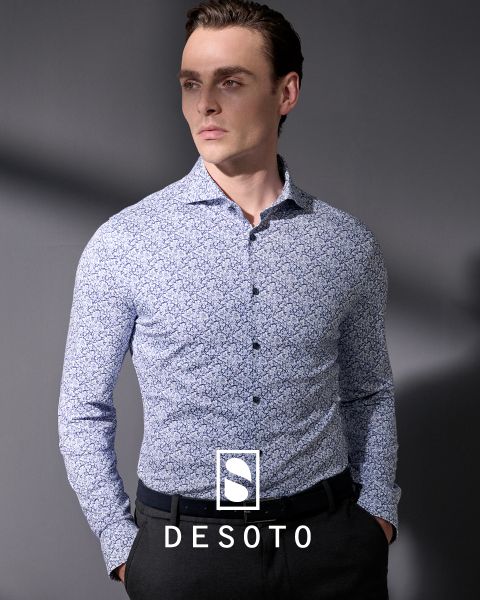 Un uomo con una camicia a maniche lunghe fantasia e pantaloni scuri è in piedi davanti a uno sfondo grigio. Guarda di lato con le mani in tasca. La scritta "DESOTO" e il logo DESOTO sono visibili nella parte inferiore.