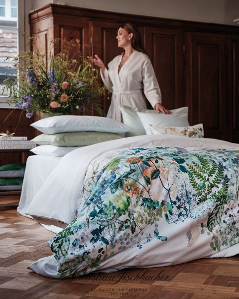 Una donna in vestaglia bianca è in piedi accanto a un letto con lenzuola a fiori. La stanza ha un pavimento in legno, un grande armadio in legno e un bouquet di fiori su un comodino. La luce del giorno entra da una finestra.