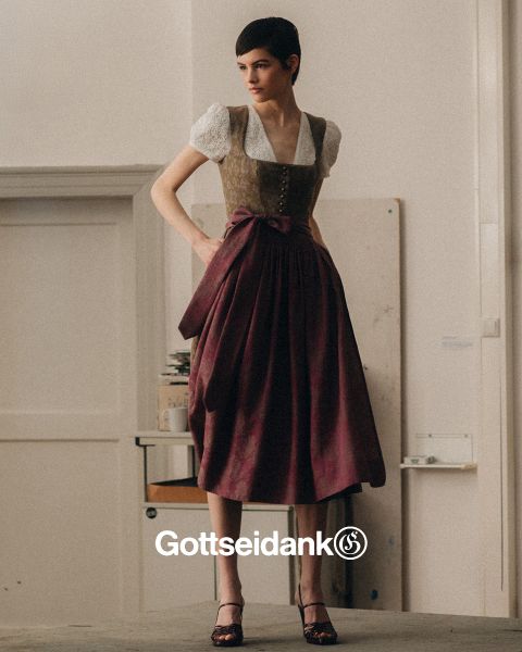 Una persona è in piedi all'interno e indossa un abito tradizionale bavarese, composto da una camicetta bianca, un gilet decorativo e una lunga gonna viola con un fiocco. Sullo sfondo si vedono una parete spoglia e una pila di libri. In basso si legge la parola "Gottseidank".