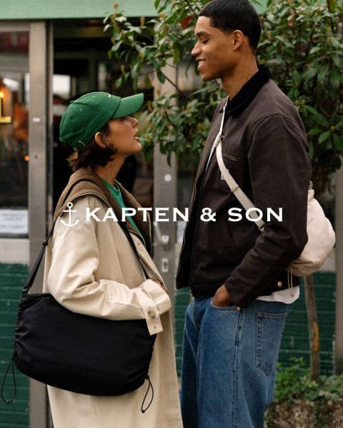 Una donna e un uomo si sorridono all'aperto. La donna indossa un cappello verde e un cappotto beige e tiene in mano una borsa nera. L'uomo indossa una giacca marrone e una borsa color crema. Al centro c'è scritto "Kapten & Son".