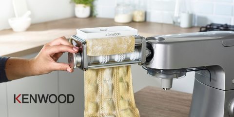 Una persona utilizza un robot da cucina Kenwood con accessorio per pasta per preparare ravioli. La pasta fresca viene stesa e tagliata in pezzi per ravioli. La cucina è arredata in stile moderno, con bicchieri e una tazza di caffè sullo sfondo.