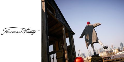 Una persona con un lungo cappotto e un cappello rosso è in equilibrio su un palo di legno vicino a un edificio rustico. Sullo sfondo si vede lo skyline della città sotto un cielo limpido. Sul lato sinistro dell'immagine si trova il logo "American Vintage".