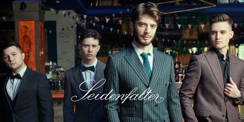 Quattro uomini in abiti formali posano con sicurezza in un ambiente elegante con decorazioni sospese. Sopra l'immagine campeggia la scritta "Seidenfalter".