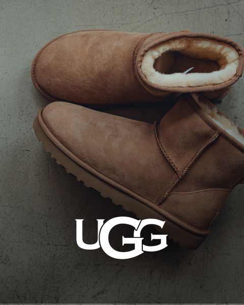 Un paio di stivali UGG in pelle scamosciata marrone con interno in peluche appoggiati su una superficie grigia. Il logo UGG è ben visibile nella parte inferiore dell'immagine.