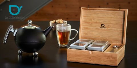 Su un tavolo scuro ci sono una teiera nera, un bicchiere di vetro pieno di tè e una scatola di legno con quattro barattoli di tè. Sulla teiera e sulla scatola si vede il logo "Bredemeijer".
