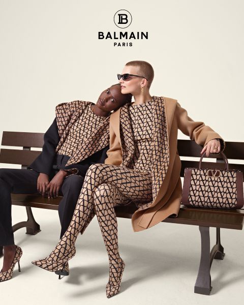 Due modelle siedono su una panchina indossando abiti Balmain coordinati con motivi geometrici. Una è appoggiata all'altra e indossa un top e pantaloni neri, mentre l'altra indossa un cappotto color cammello e occhiali da sole. Accanto a loro c'è una borsa a motivi. In alto, il logo Balmain.