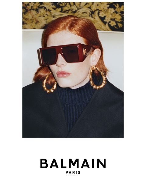 Una persona con capelli rossi, un paio di occhiali da sole scuri oversize, grandi orecchini dorati e un top scuro a collo alto posa davanti a uno sfondo a motivi. L'immagine mostra il logo BALMAIN Paris nella parte inferiore.