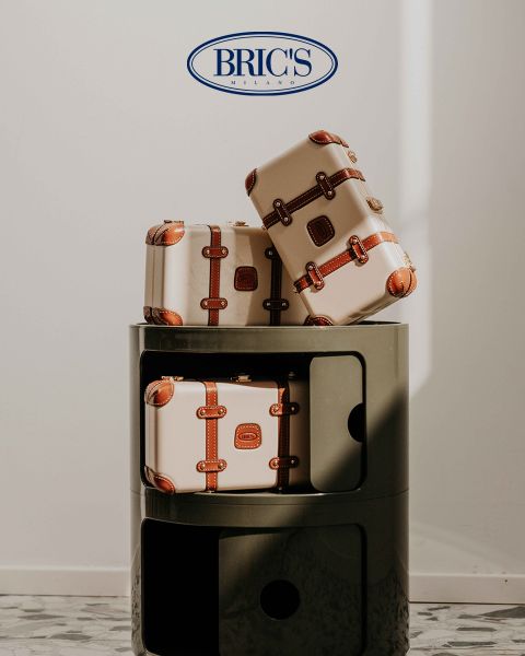 Tre valigie beige con cinturini in pelle marrone sono impilate su un moderno scaffale rotondo grigio scuro. Sopra, il logo BRIC'S MILANO spicca su una parete bianca. La luce del sole proietta ombre delicate sulla scena.