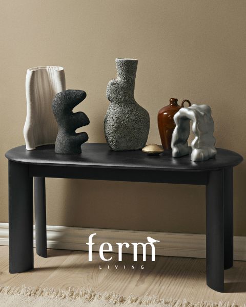Un tavolo nero con sculture astratte in ceramica di varie forme e texture si trova davanti a una parete beige. Il pavimento è in legno chiaro e in basso si vede il logo "ferm LIVING".