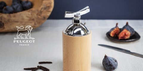 Un macinapepe in legno e acciaio inossidabile si trova accanto a fichi interi e tagliati su una tovaglia. Accanto c'è un coltello. Nell'angolo in alto a sinistra si vede il logo Peugeot con un leone. Sullo sfondo si vede una ciotola di legno con capsule di spezie.