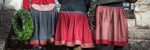 Tre persone vestite a festa sono in piedi una accanto all'altra e tengono in mano una ghirlanda verde. Indossano gonne a motivi nei toni del rosso e del grigio scuro, giacche calde e guanti. La scena sembra svolgersi all'aperto su gradini di pietra.