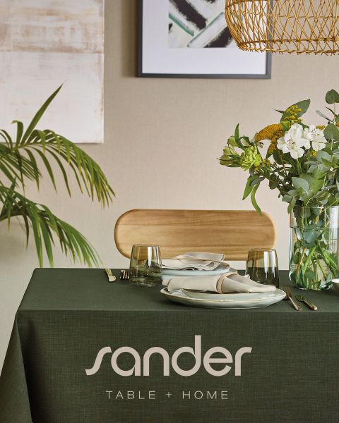 Un tavolo da pranzo moderno con una tovaglia verde, piatti, bicchieri, tovaglioli, una composizione floreale e la scritta „sander TABLE + HOME“.