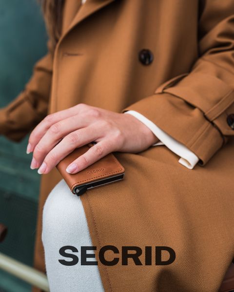 Una persona con un cappotto marrone tiene un piccolo portafoglio SECRID marrone sul ginocchio. Sul bordo inferiore dell'immagine è stampata la parola "SECRID" in grandi lettere.