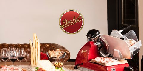 Su un tavolo con vari tipi di carne, formaggio, grissini e bicchieri di vino, si trova un'affettatrice antica per carne rossa. Sullo sfondo si intravedono un divano in pelle e un logo "Berkel". L'ambiente è accogliente e rustico.