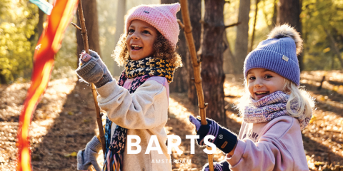 Due bambini corrono attraverso una foresta soleggiata. Indossano cappelli invernali, sciarpe e guanti. Sorridono e tengono in mano bastoni con nastri colorati. In basso nell'immagine si vede il testo "BARTS AMSTERDAM".