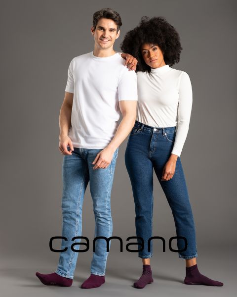 Un uomo in maglietta bianca e jeans e una donna in top bianco a maniche lunghe e jeans posano abbracciati. Entrambi sono a piedi nudi. In basso nell'immagine c'è la parola "camano".