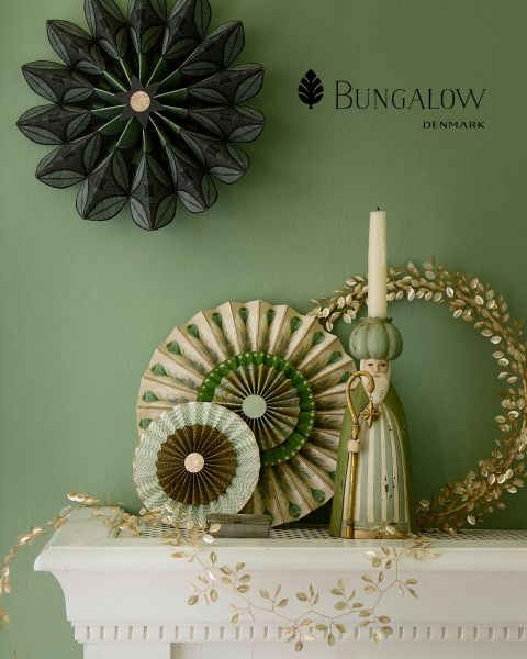 Una parete verde è decorata con oggetti decorativi: un ornamento nero a forma di stella, ventagli di carta rotondi in verde e beige, una figura rustica di una persona in una veste con un cappello alto, una ghirlanda decorativa dorata e un camino bianco con motivi floreali. Il testo recita "Bungalow Danimarca".