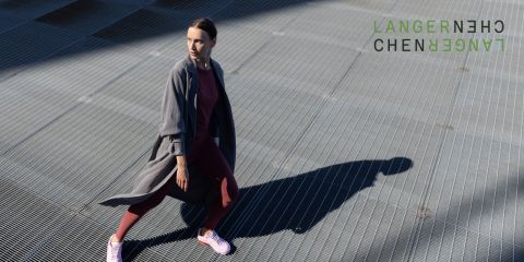 Una persona in abbigliamento sportivo e un lungo cappotto cammina su una superficie a griglia metallica, proiettando un'ombra. Nell'angolo in alto a destra si vede il testo astratto verde "LANGERNECHE".