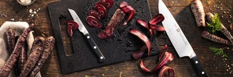 Un tavolo di legno scuro con un tagliere nero su cui sono appoggiati due coltelli e carote viola tagliate. Carote intere e foglie di carota sono sparse intorno, creando una scena di cucina rustica.