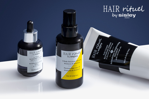 Una collezione di prodotti per la cura dei capelli del marchio "HAIR rituel by sisley" su una superficie. Il set include un flacone contagocce, un flacone spray e un tubetto, tutti con la scritta "cura dei capelli e del cuoio capelluto" e uno sfondo scuro.
