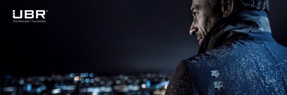 Una persona con una giacca guarda sopra la spalla un paesaggio urbano notturno. Sulla giacca ci sono gocce d'acqua che riflettono le luci della città. Nell'angolo in alto a sinistra si vedono il logo UBR e le parole "Technology Tailoring".